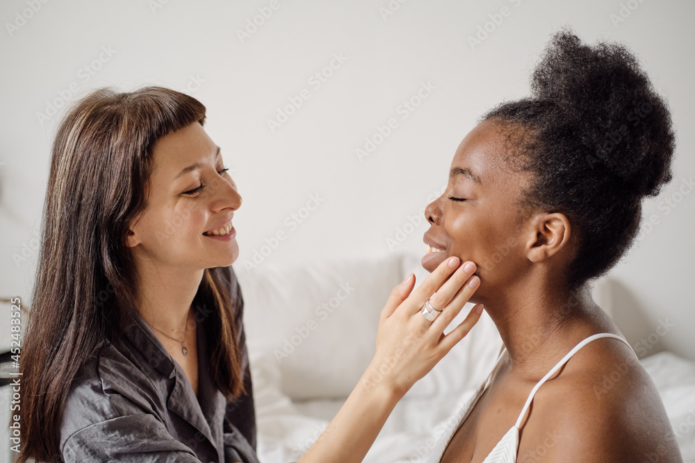 Happy young woman applying cream on face of her African girlfriend Happy young woman applying cream on face of her African girlfriend