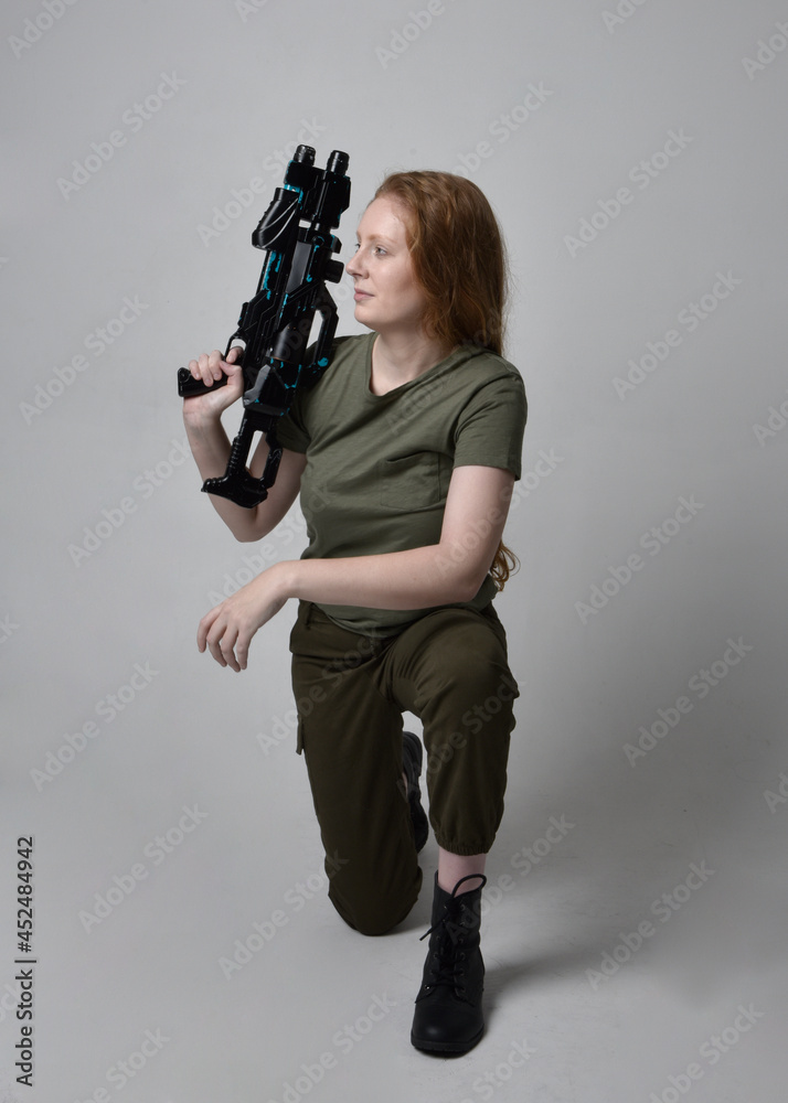 Full length portrait of pretty red haired woman wearing army green khaki shirt, utilitarian pants and boots. Holding a gun as weapon, isolated on studio background. Full length portrait of pretty red haired woman wearing army green khaki shirt, utilitarian pants and boots. Holding a gun as weapon, isolated on studio background.