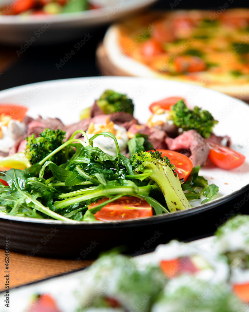 Healthy salad made from meat, cherry tomatoes, broccoli, goat cheese and fresh arugula served in a white plate.