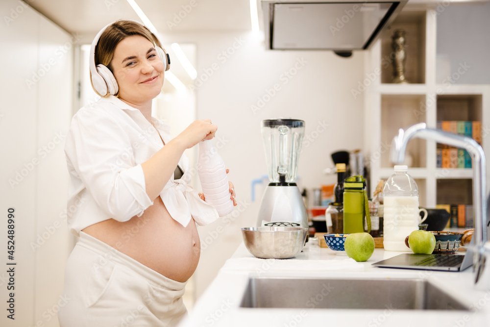 White pregnant woman in headphones making smoothy at home White pregnant woman in headphones making smoothy at home