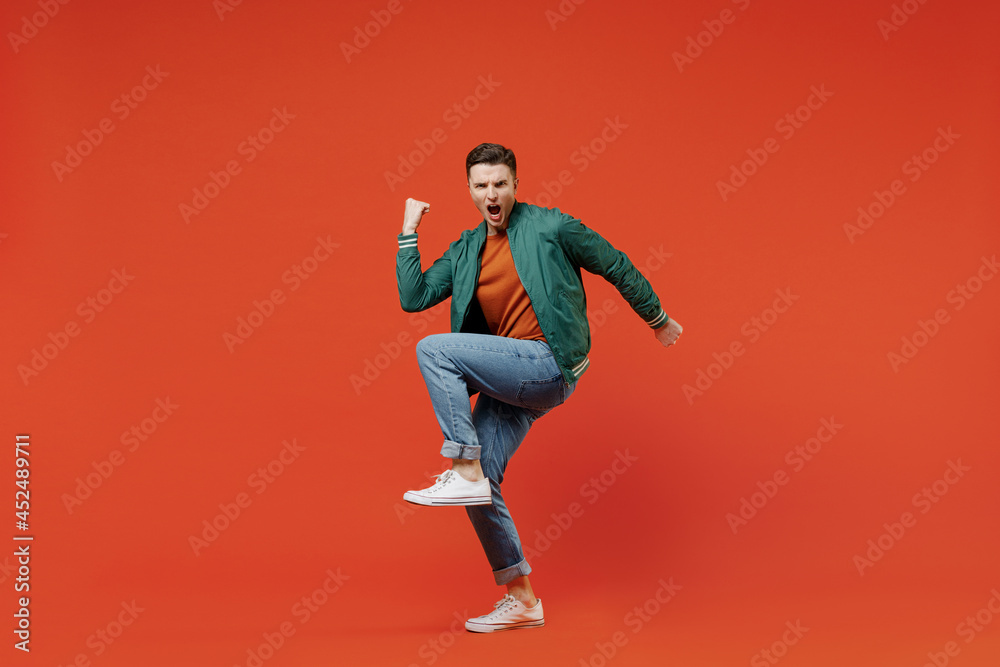 Full size body length smiling vivid young brunet man 20s wears red t-shirt green jacket doing winner gesture celebrating clenching fists say yes isolated on plain orange background studio portrait