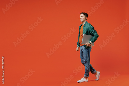 Full size body length side view profile young brunet man 20s wear red t-shirt green jacket hold laptop pc under his hand move look ahead computer isolated on plain orange background studio portrait