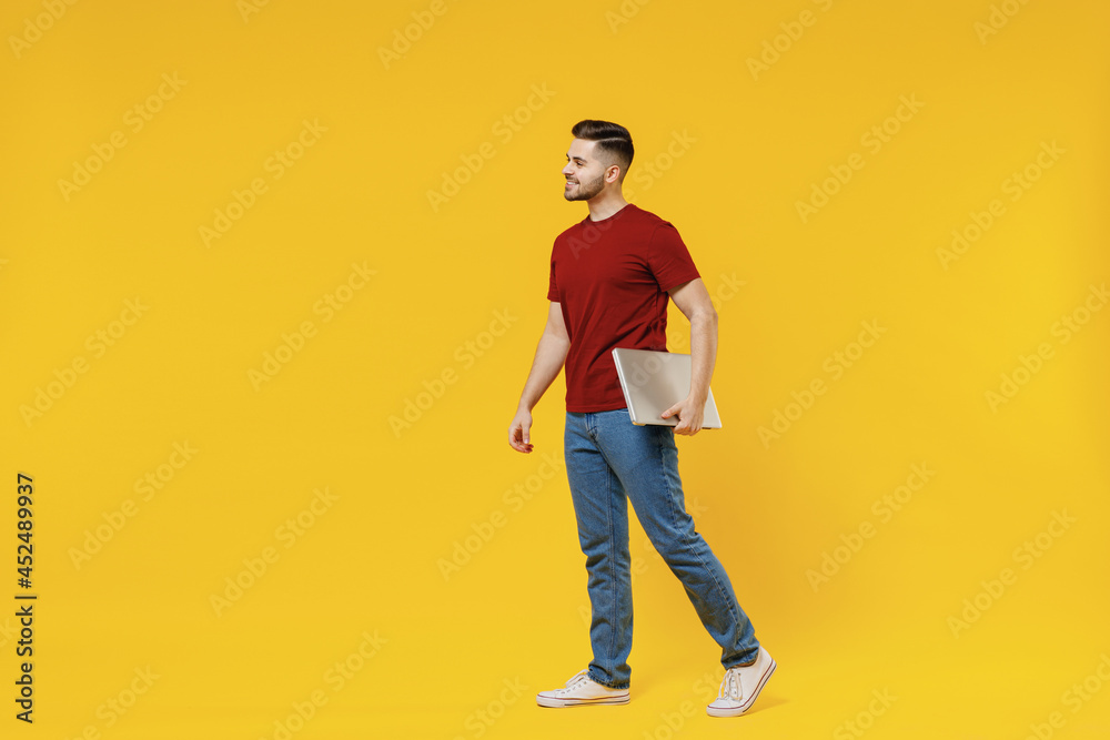 Full length smiling happy young man in red tshirt casual clothes hold closed laptop pc computer work look aside isolated on plain yellow color wall background studio portrait People lifestyle concept Full length smiling happy young man in red tshirt casual clothes hold closed laptop pc computer work look aside isolated on plain yellow color wall background studio portrait People lifestyle concept