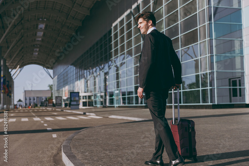 Full size back side view young traveler businessman man in black dinner suit walking going outside at international airport terminal with suitcase valise Air flight business trip lifestyle concept © ViDi Studio