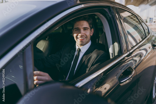 Side profile view young driver confident smiling businessman man 20s wear black dinner suit driving car taxi hold steering wheel look camera Vehicle transport traffic lifestyle business trip concept © ViDi Studio