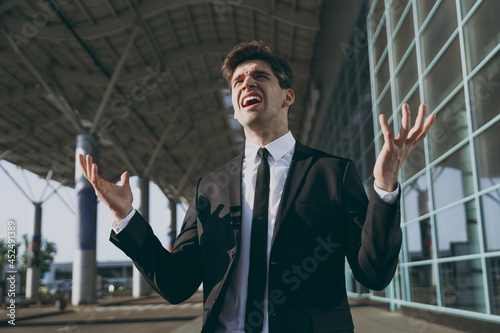 Dissatisfied sad indignant young traveler businessman man wear black classic tie suit standing outside at international airport terminal spread hands. People air flight business trip lifestyle concept