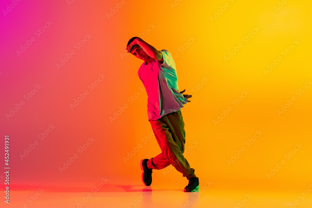 Full length portrait of young stylish man, break dancing dancer in casual clothes isolated over gradient pink yellow background at dance hall in neon light.