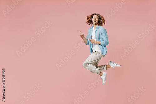 Full length young happy fun cool man 20s with long curly hair wear blue shirt white t-shirt point index finger aside on workspace area jump high isolated on pastel plain pink wall background studio