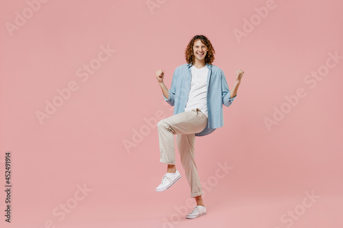 Full length young excited happy man with long curly hair wear blue shirt white t-shirt do winner gesture with raised up leg camera isolated on pastel plain pink color wall background studio portrait