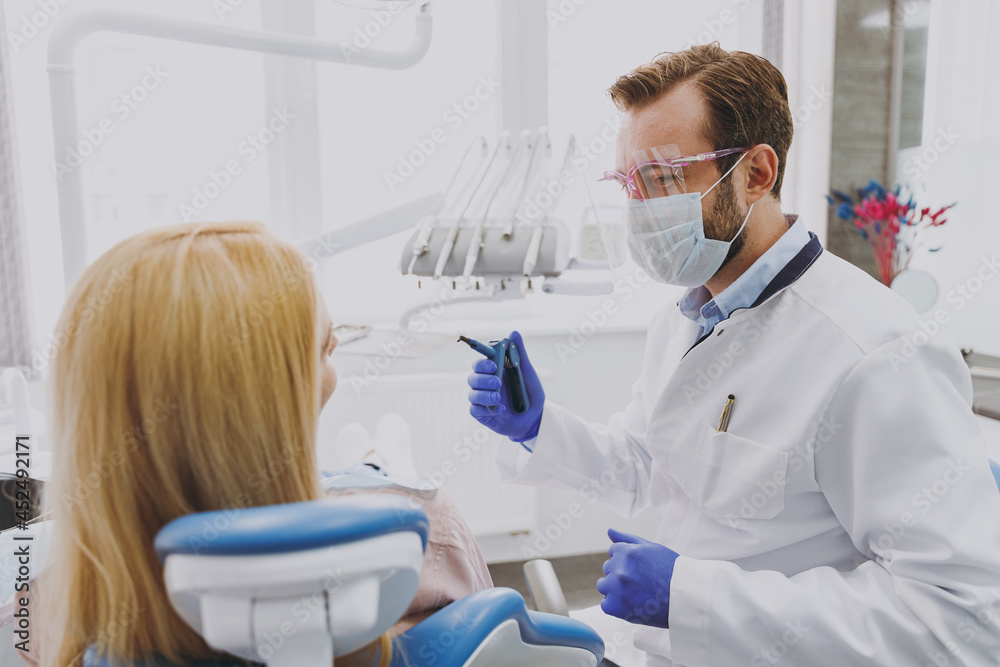 Young smiling man doctor in face mask shields and googles examine mouth cavity of patient woman sit at dentist office chair indoor cabinet near stomatologist Healthcare enamel caries decay treatment Young smiling man doctor in face mask shields and googles examine mouth cavity of patient woman sit at dentist office chair indoor cabinet near stomatologist Healthcare enamel caries decay treatment