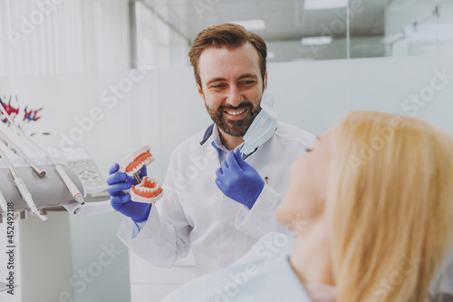 Young fun orthodontist man wear gown doctor dentist show artificial plastic jaw bite for patient woman sitting at dentist office chair indoor cabinet near stomatologist. Healthcare enamel treatment