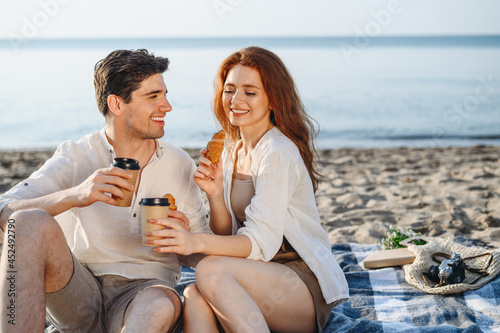 Young fun couple family two man woman in white clothes hug sitting sand plaid have picnic eat croissant drink coffee rest together at sunrise over sea beach ocean outdoor seaside in summer day sunset. © ViDi Studio