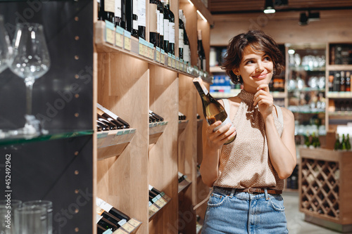Young minded woman 20s wear casual clothes shopping at supermaket grocery store buy choosing white wine alcohol hold bottle prop up chin inside hypermarket People purchasing gastronomy food concept