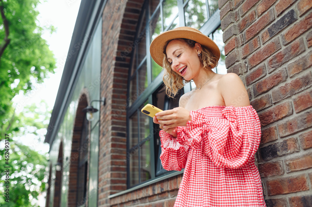 Bottom side view young smiling happy caucasian woman in pink dress hat use mobile cell phone walk in city center stand outdoor near town brick old building People urban summer time lifestyle concept.