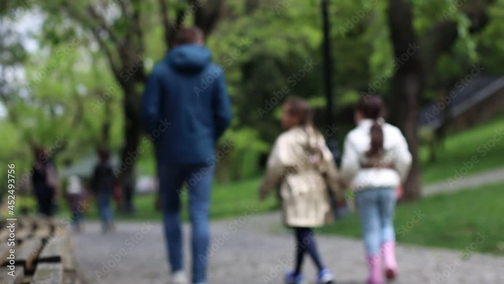 Back view of happy family people enjoying walking on street in city park. Dad walks with daughters. Slow motion. Blurred background Back view of happy family people enjoying walking on street in city park. Dad walks with daughters. Slow motion. Blurred background