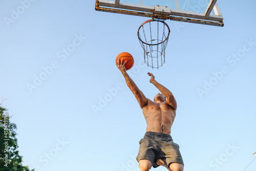 Bottom view young muscular sporty sportsman man naked torso shoot free throw jump high train hold in hand ball at basketball game playground court on sky background Outdoor courtyard sports concept. © ViDi Studio