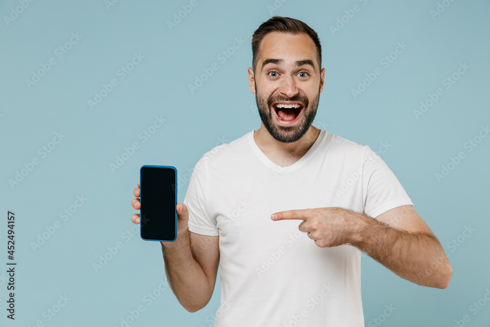 Young surprised man in blank print design white t-shirt point index finger on mobile cell phone with blank screen workspace area isolated on plain pastel light blue color background studio portrait Young surprised man in blank print design white t-shirt point index finger on mobile cell phone with blank screen workspace area isolated on plain pastel light blue color background studio portrait