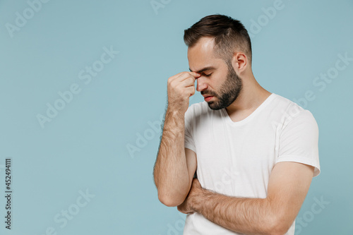 Fotografie Young sad displeased tired sick man 20s wear casual white t-shirt keep eyes closed rub put hand on nose isolated on plain pastel light blue color background studio portrait