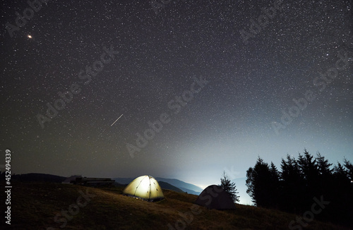 Night camping in the mountains. Two tents  one of which illuminated  set up on mountain meadow. Panoramic view of evening sky with multiple stars.