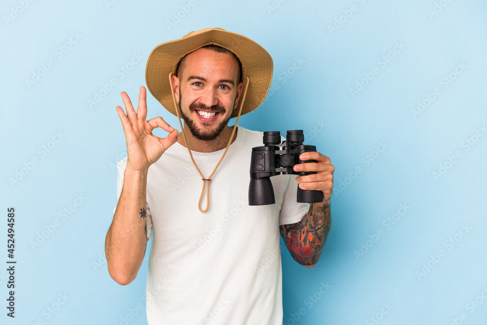 Young caucasian man with tattoos holding binoculars isolated on blue background cheerful and confident showing ok gesture. Young caucasian man with tattoos holding binoculars isolated on blue background cheerful and confident showing ok gesture.