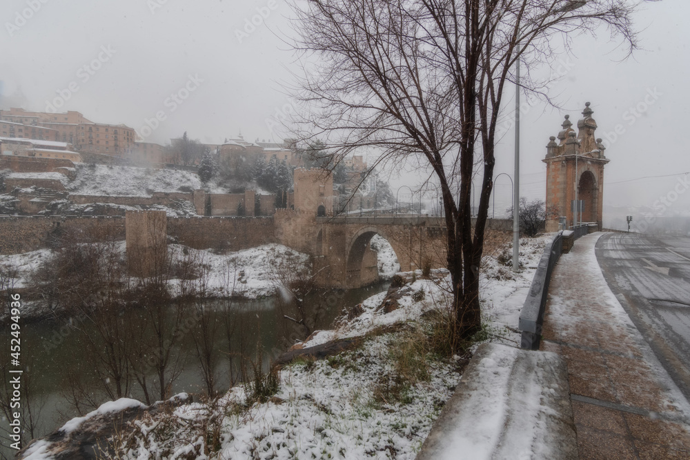 City landscape of snowy old city and castle with clouds and fog; View of the Tajo river, Alcázar and Alcántara bridge from Toledo during the storm Filomena, World Heritage Site, Spain. Horizontal view