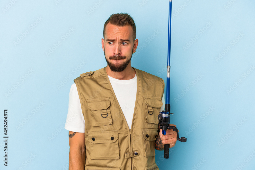 Young caucasian fisherman with tattoos holding rod isolated on blue background confused, feels doubtful and unsure. Young caucasian fisherman with tattoos holding rod isolated on blue background confused, feels doubtful and unsure.