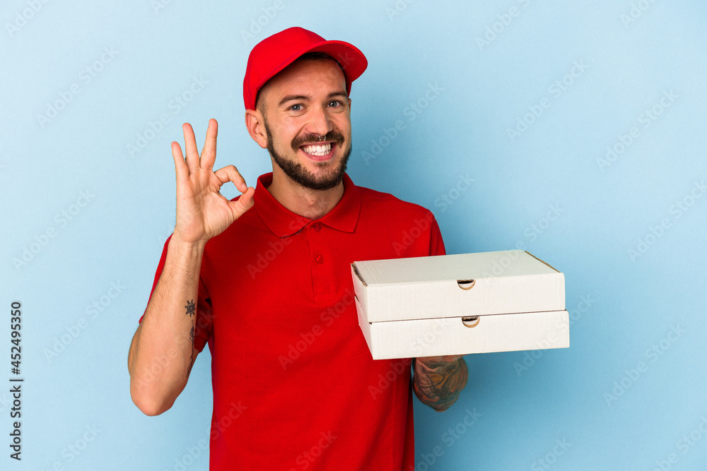 Young caucasian delivery man with tattoos holding pizzas isolated on blue background cheerful and confident showing ok gesture. Young caucasian delivery man with tattoos holding pizzas isolated on blue background cheerful and confident showing ok gesture.