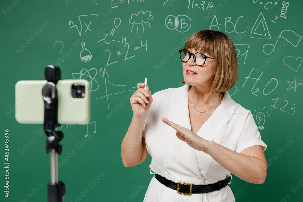 Smart teacher mature elderly lady woman 55 wear shirt glasses work with mobile cell phone on tripod on lockdown quarantine explain new material isolated green wall chalk blackboard background studio.