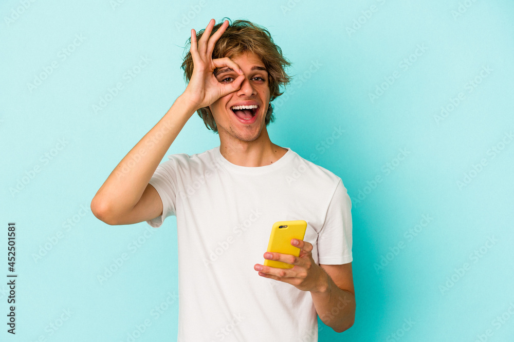 Young caucasian man with makeup holding mobile phone isolated on blue background excited keeping ok gesture on eye. Young caucasian man with makeup holding mobile phone isolated on blue background excited keeping ok gesture on eye.