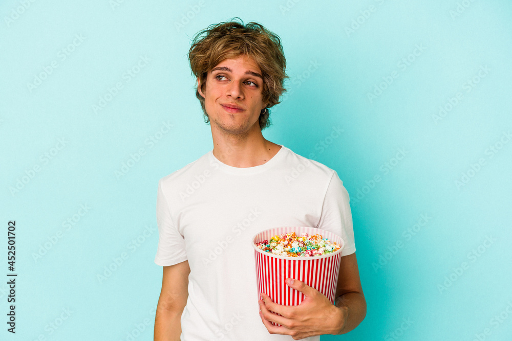 Young caucasian man with makeup holding popcorn isolated on blue background  dreaming of achieving goals and purposes