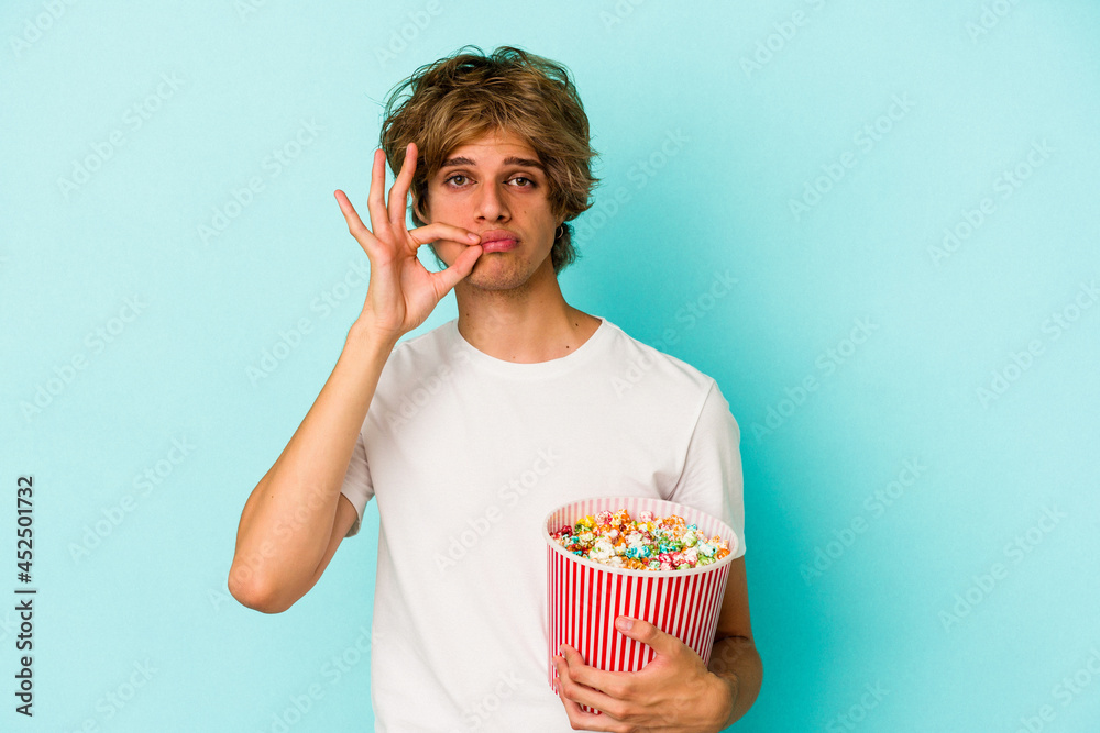 Young caucasian man with makeup holding popcorn isolated on blue background  with fingers on lips keeping a secret.