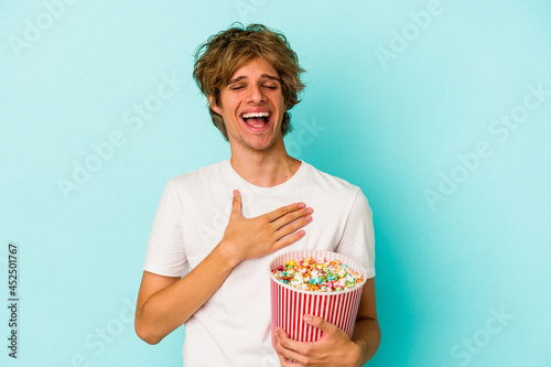 Young caucasian man with makeup holding popcorn isolated on blue background laughs out loudly keeping hand on chest.