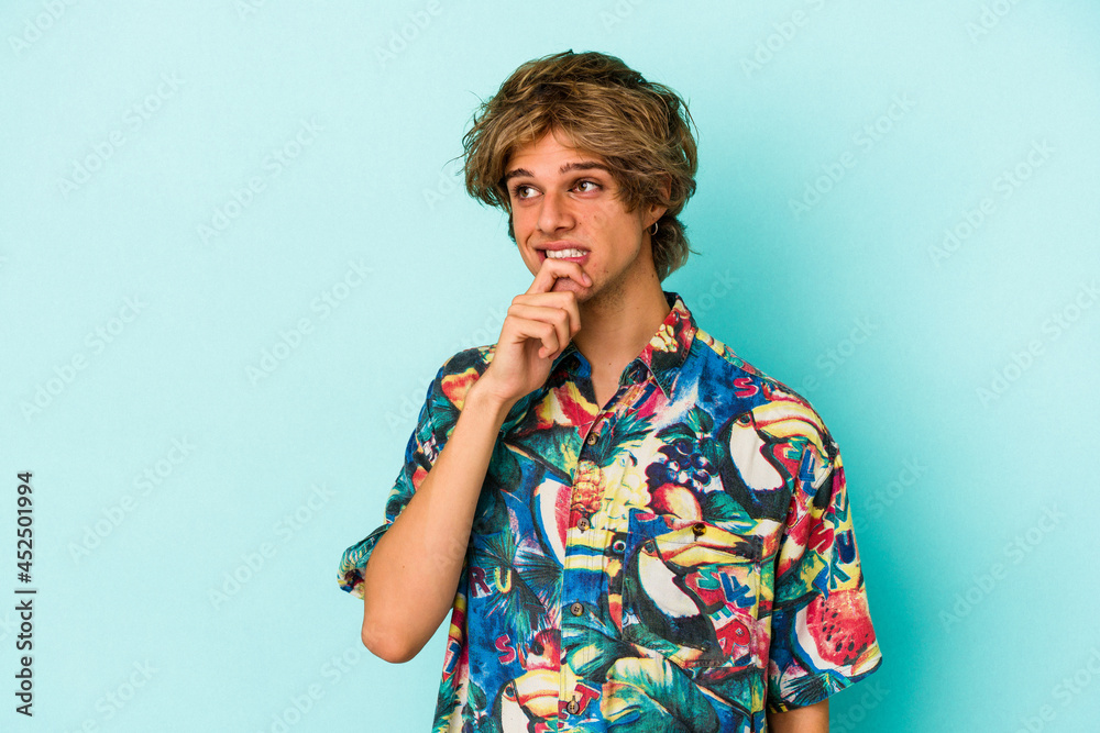 Young caucasian man with makeup wearing a summer clothes isolated on blue background relaxed thinking about something looking at a copy space. Young caucasian man with makeup wearing a summer clothes isolated on blue background relaxed thinking about something looking at a copy space.