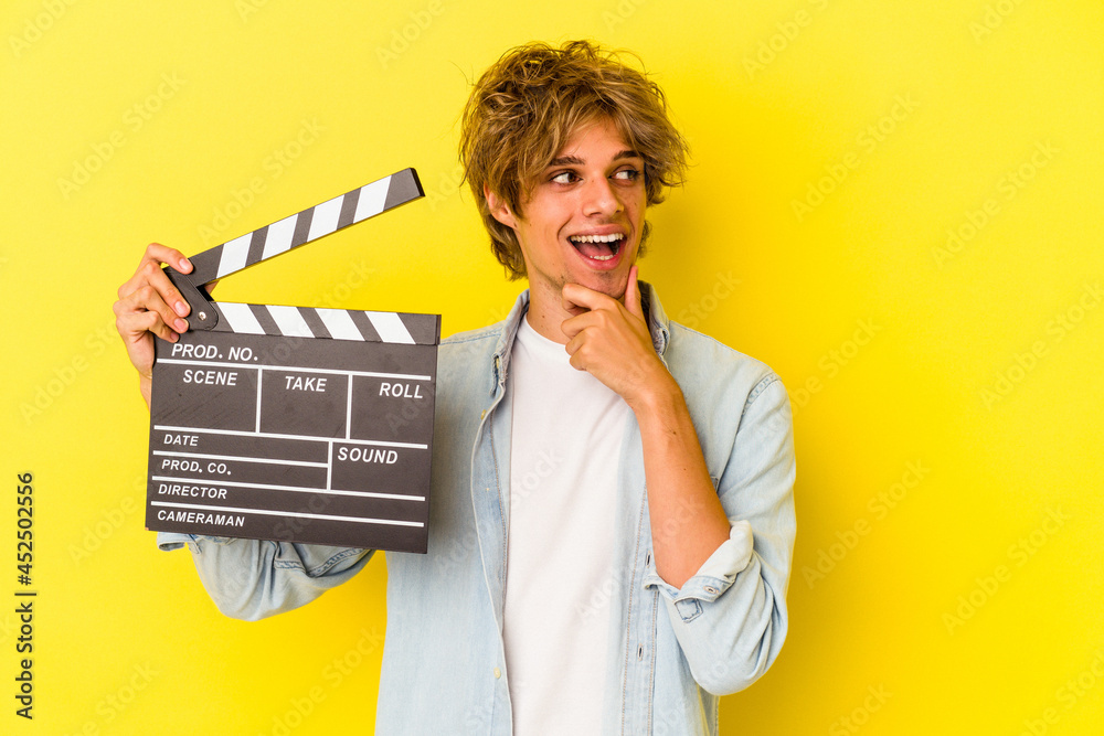 Young caucasian man with makeup holding clapperboard isolated on yellow background  relaxed thinking about something looking at a copy space.