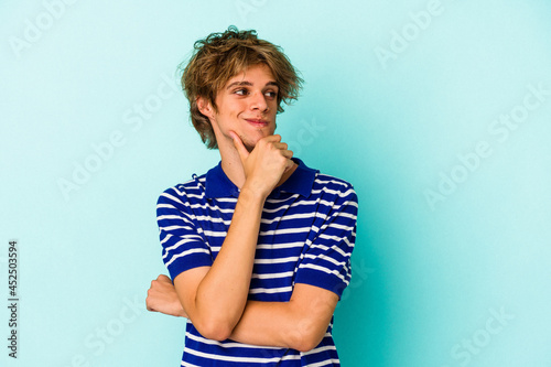 Young caucasian man with make up isolated on blue background looking sideways with doubtful and skeptical expression.