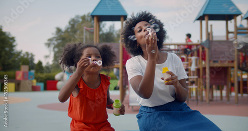 Afro-american mother and daughter play soap bubbles at playground.