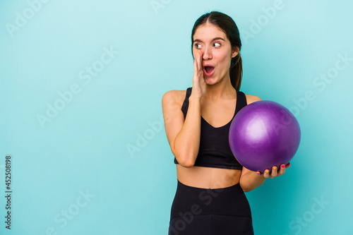 Young caucasian fitness woman holding a ball isolated on blue background is saying a secret hot braking news and looking aside