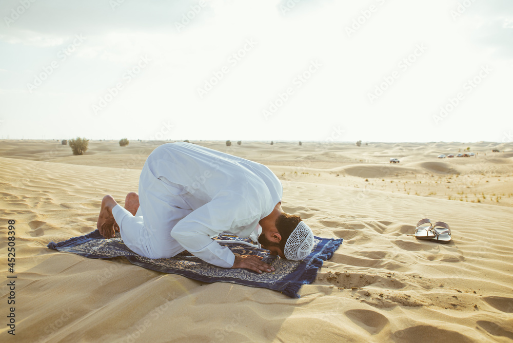 Man with white traditional kandura from uae praying in the desert on the carpet
