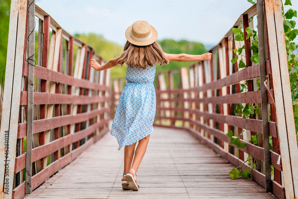 Rear view of a little girl in a hat in the middle of a wooden bridge in a green summer park. The concept of freedom and a happy childhood