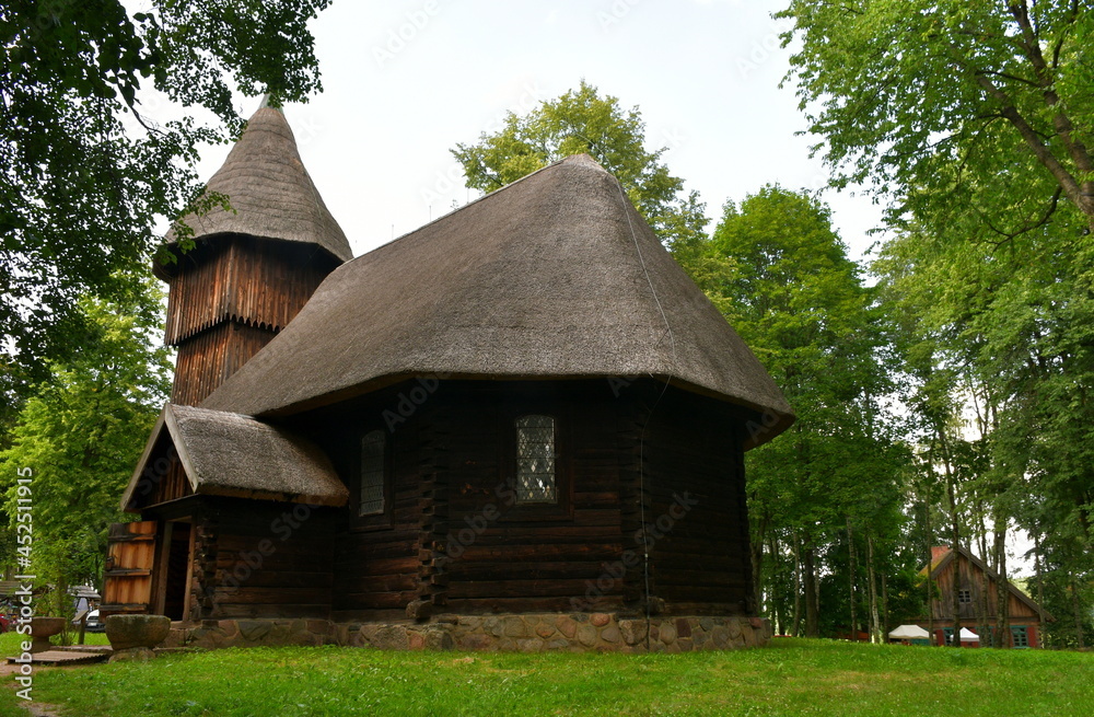 A close up on an old abandoned wooden church with a thatch angled roof, glass windows, and a tall tower standing in the middle of a forest or moor near a Polish village or cottage in summer