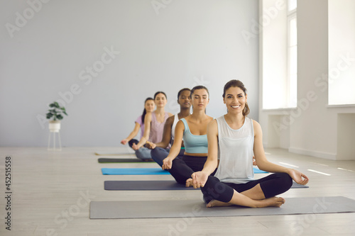 Calm relaxed beautiful young women having yoga class at gym. Group of multiracial ladies doing easy asanas and breathing exercises together with happy smiley yoga trainer. Healthy lifestyle concept