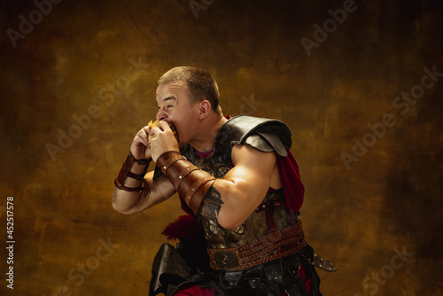 Portrait of young man, medieval warrior or knight in war equipment eating burger isolated on vintage dark background. Comparison of eras, art, history, humor, retro style concept photo