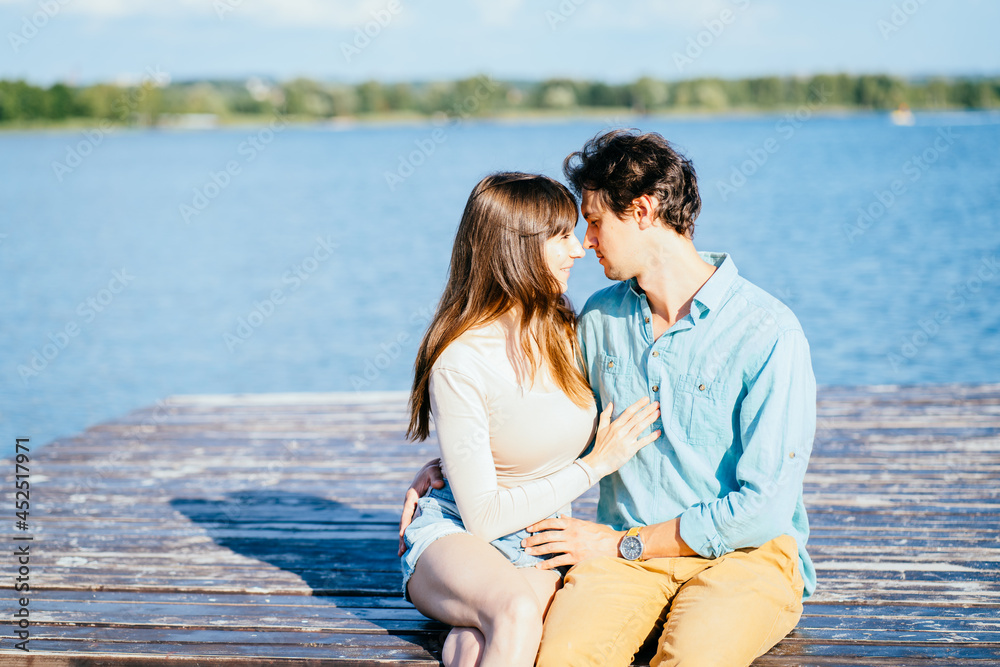 Couple of two, man and woman, sitting together on pier of wooden bridge in the sea or lake and hugging. Sunny joyful summer day or evening concept.