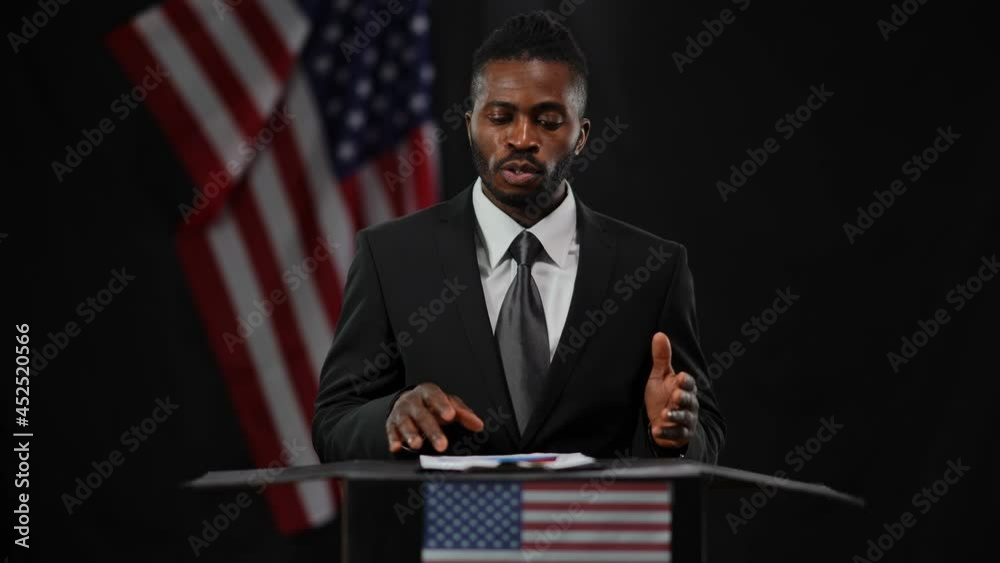 Serious African American man in elegant formal black suit speaking gesturing standing at tribune with USA flag at background. Portrait of confident speaker talking with audience