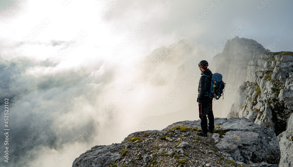 Man with backpack high above the misty mountain valley. Lone person  looks onwards at a mountain shrouded in mist and clouds with the peak visible. Scenic landscape photo composite.