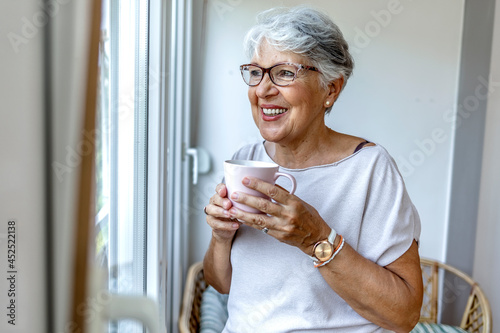 Shot of an elderly woman sitting by the window holding a cup. An old woman wearing eyeglasses standing inside the house, holding a cup of coffee. Smiling senior woman holding cup of coffee at home.