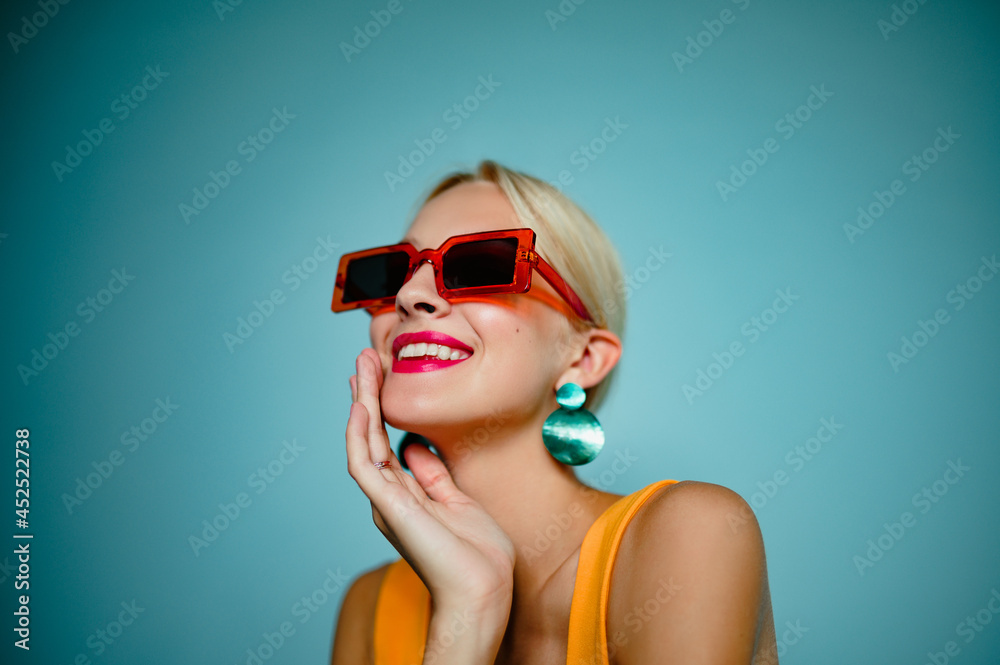 Φωτογραφία Happy smiling fashionable woman wearing trendy summer orange sunglasses, green shell earrings, posing on blue background