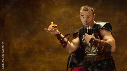 Photography Portrait of young man, medieval warrior or knight in war equipment eating burger isolated on vintage dark background