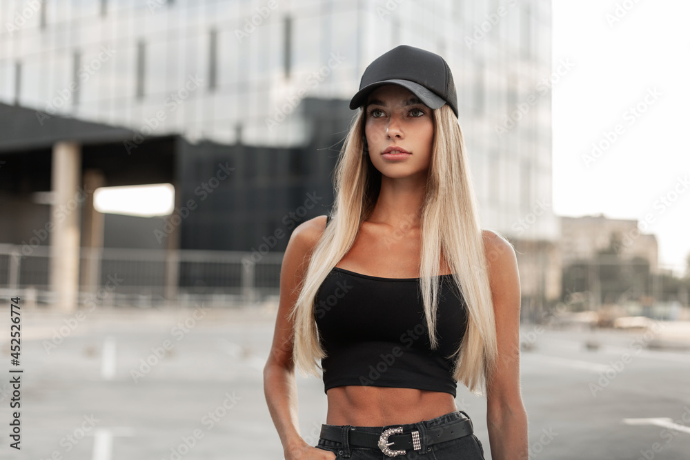 Pretty model hipster woman in a black baseball cap with a black stylish tank top and denim shorts with a belt stands in the city. Modern urban female style look outfit
