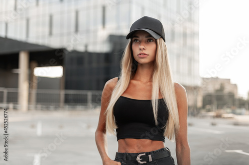 Pretty model hipster woman in a black baseball cap with a black stylish tank top and denim shorts with a belt stands in the city. Modern urban female style look outfit photo